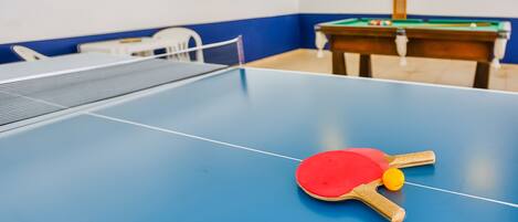 Una mesa de ping pong con una paleta roja y una pelota amarilla en primer plano, una mesa de billar al fondo y una silla blanca a la izquierda.