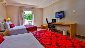 Habitación de hotel con dos camas, un escritorio, una silla, un televisor y una ventana con vista a la vegetación.