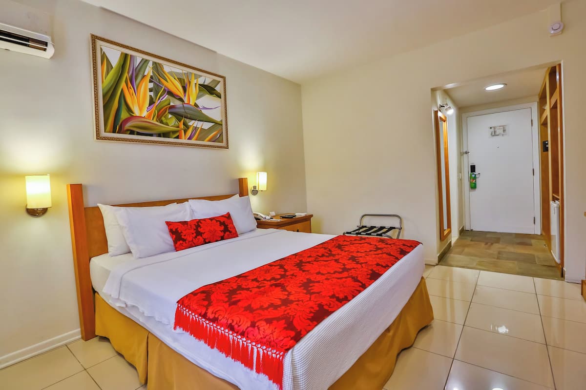 A hotel room with a bed, bedside lamps, a nightstand, a door leading to another room, and a wall-mounted air conditioner.