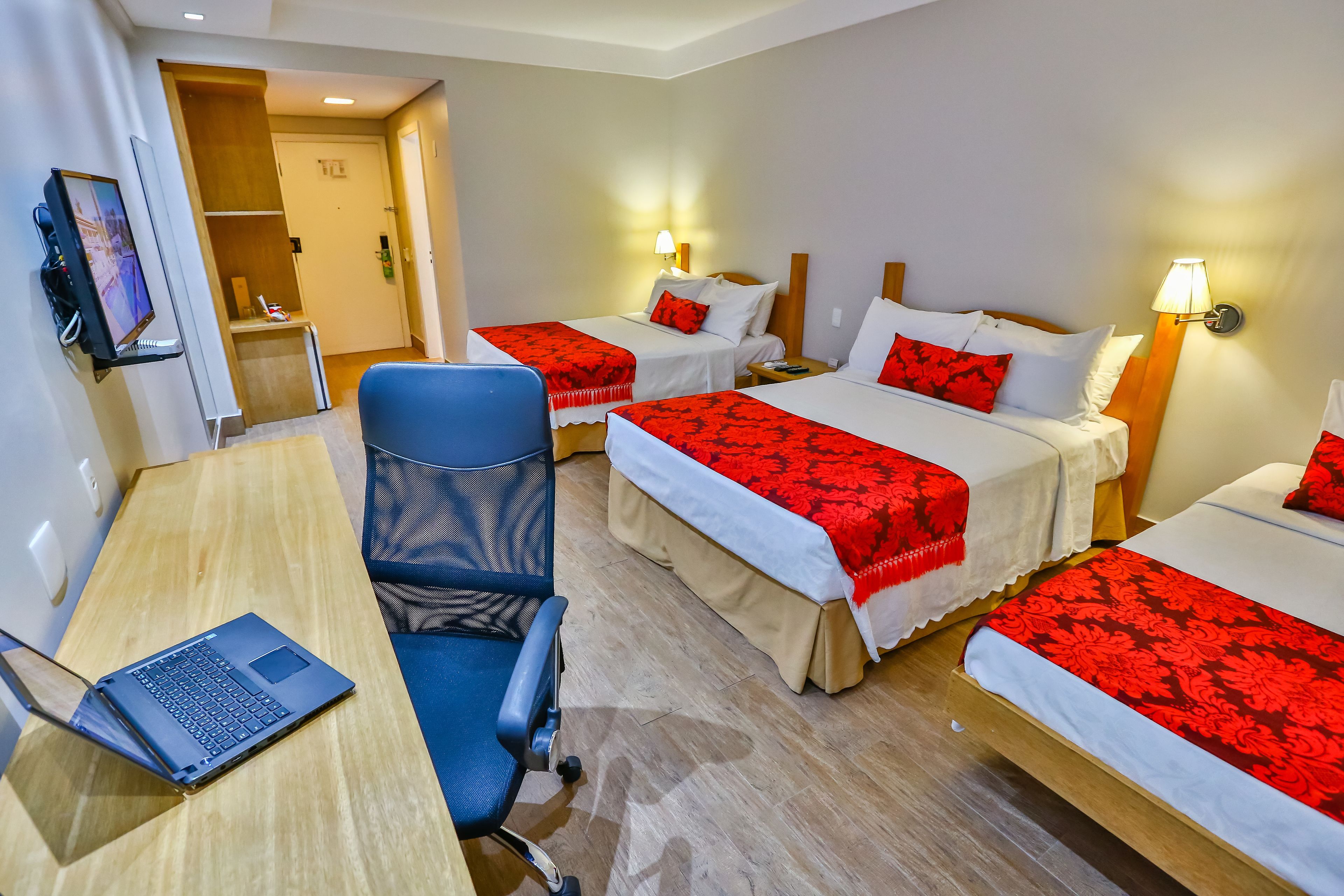 Quarto de hotel com duas camas, uma escrivaninha com um laptop, uma cadeira e uma televisão.