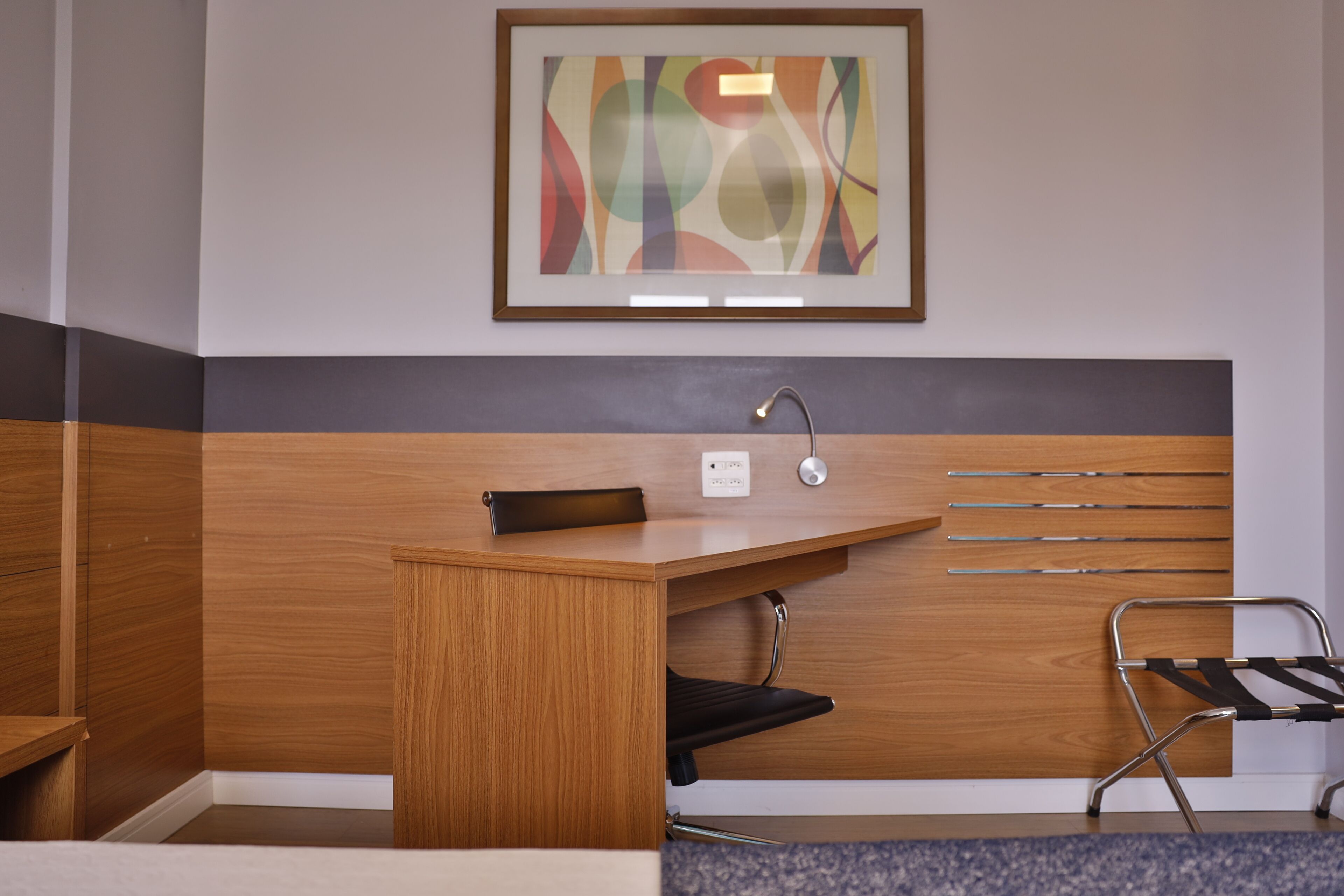 A hotel room with a wooden desk, a black chair, a framed abstract painting, and a wall-mounted towel rack.