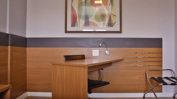 A hotel room with a wooden desk, a black chair, a framed abstract painting, and a wall-mounted towel rack.