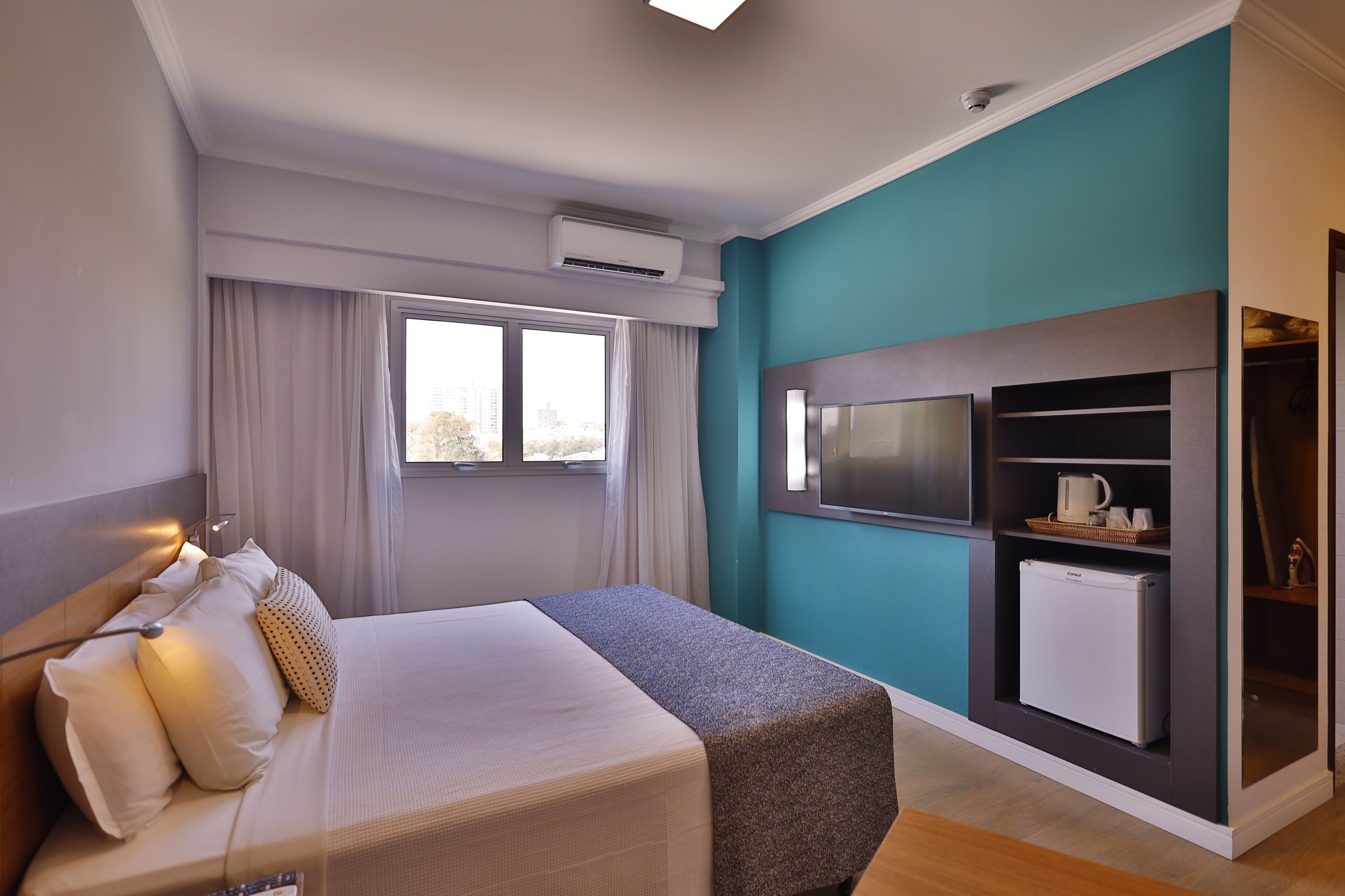 A modern hotel room with a bed, a TV, a mini-fridge, and a view of the city through the window.