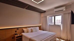 Una habitación de hotel moderna con una cama grande, cabecera de madera, mesitas de noche integradas y vistas a la ciudad a través de amplias ventanas.