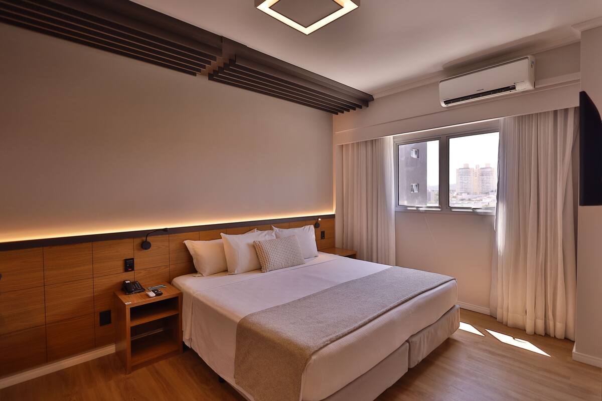 Una habitación de hotel moderna con una cama grande, cabecera de madera, mesitas de noche integradas y vistas a la ciudad a través de amplias ventanas.
