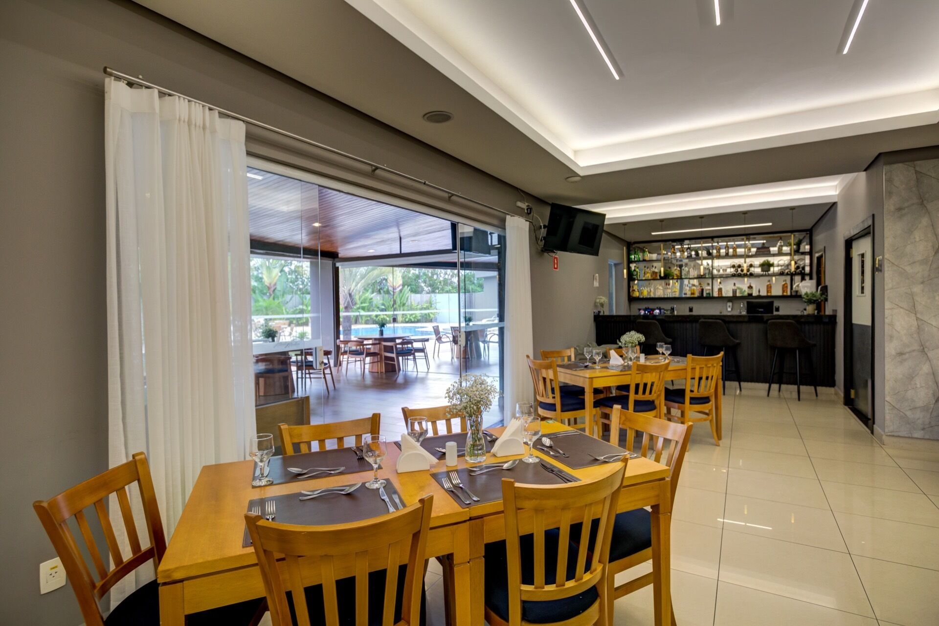 A dining area with wooden tables and chairs, a bar with shelves of bottles, and a large window offering a view of the outdoors.