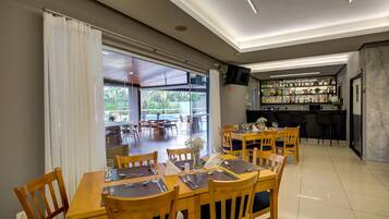 A dining area with wooden tables and chairs, a bar with shelves of bottles, and a large window offering a view of the outdoors.