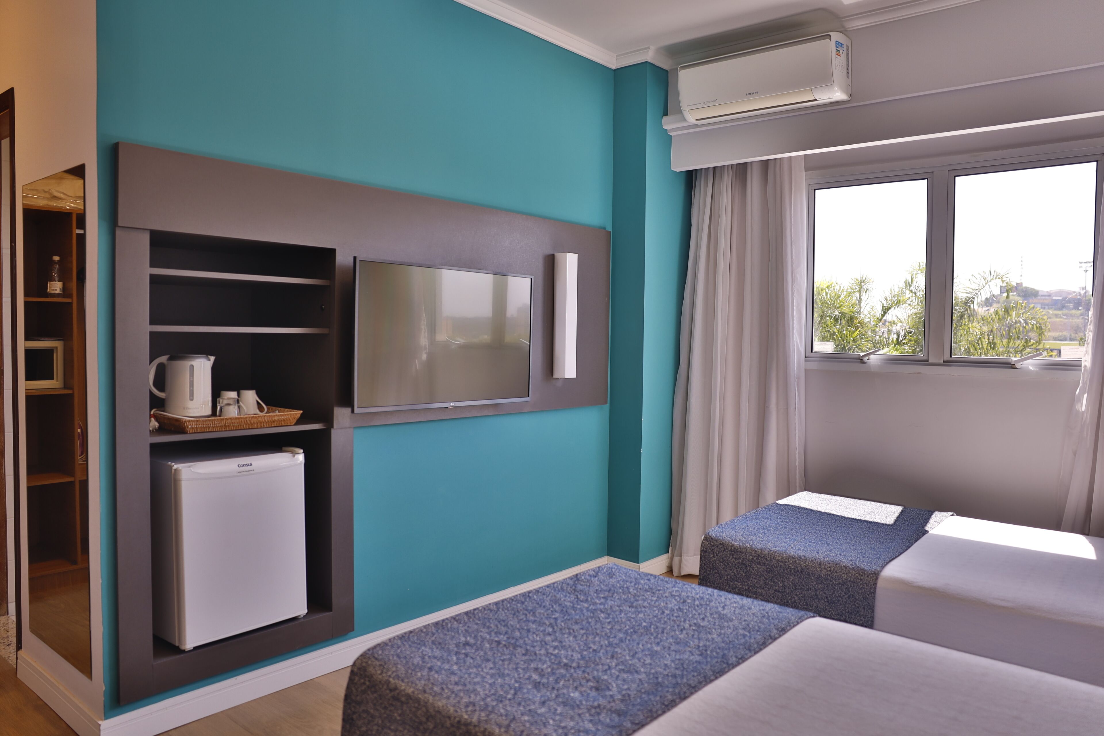 A hotel room with teal walls, a TV mounted on the wall, a mini-fridge, a microwave, a window with curtains, and two beds.