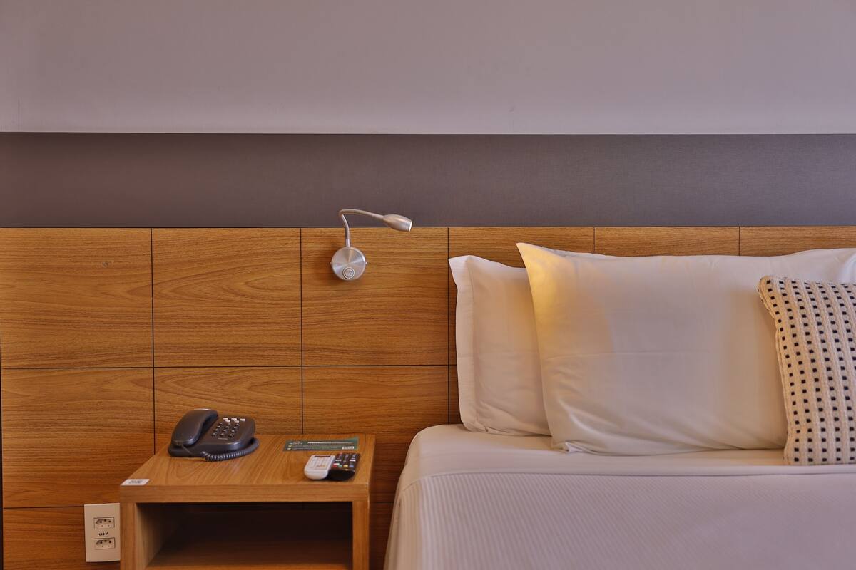 Habitación de hotel con cabecera de madera, mesita de noche con teléfono, control remoto y toma de corriente.