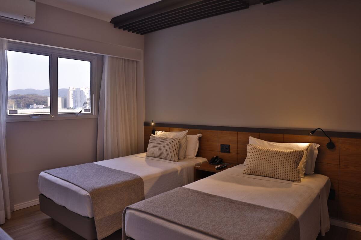 Una habitación de hotel con dos camas individuales, una mesita de noche con teléfono y una ventana con vistas a edificios y colinas.