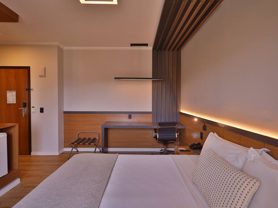 A modern hotel room with a bed, a desk, and a chair. There is a wooden door and a small refrigerator.