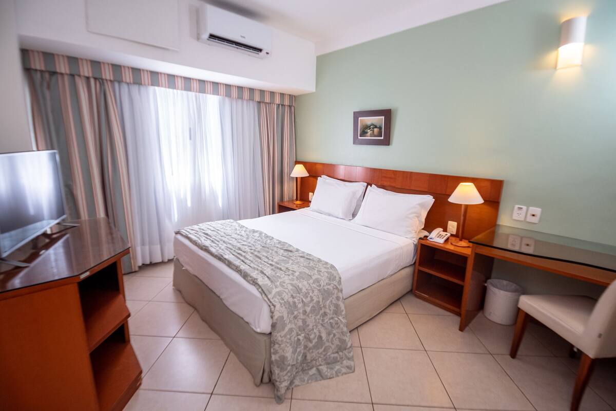 A hotel room with a bed, bedside lamps, a desk, a chair, a TV, and a picture on the wall.