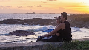 Una pareja sentada sobre un borde de piedra con vista al océano al atardecer.