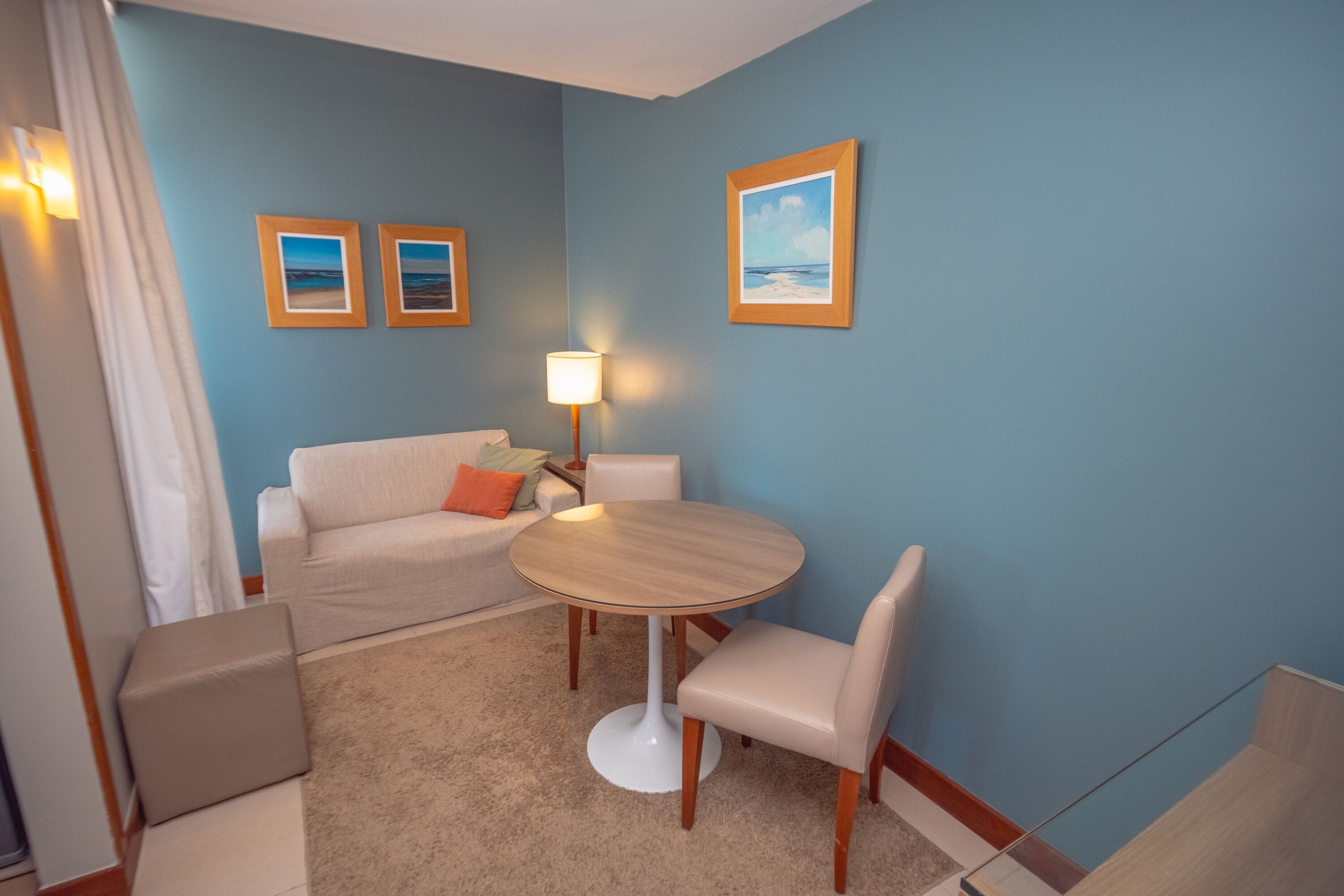 Habitación de hotel con una mesa redonda, dos sillas, un sofá y una lámpara. Hay tres cuadros enmarcados en la pared.
