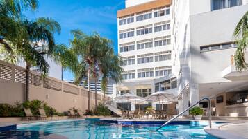 A swimming pool with lounge chairs and umbrellas, surrounded by palm trees and a modern building with multiple floors.