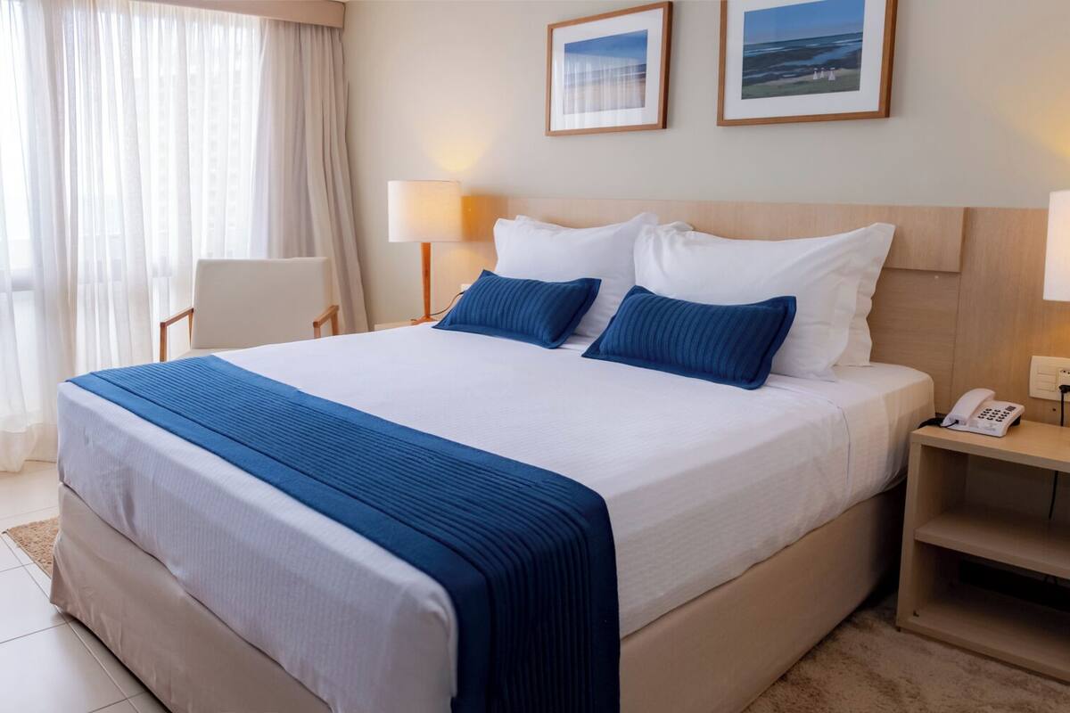 A hotel room with a large bed, two bedside lamps, a phone on the nightstand, and a view of the sea through the window.