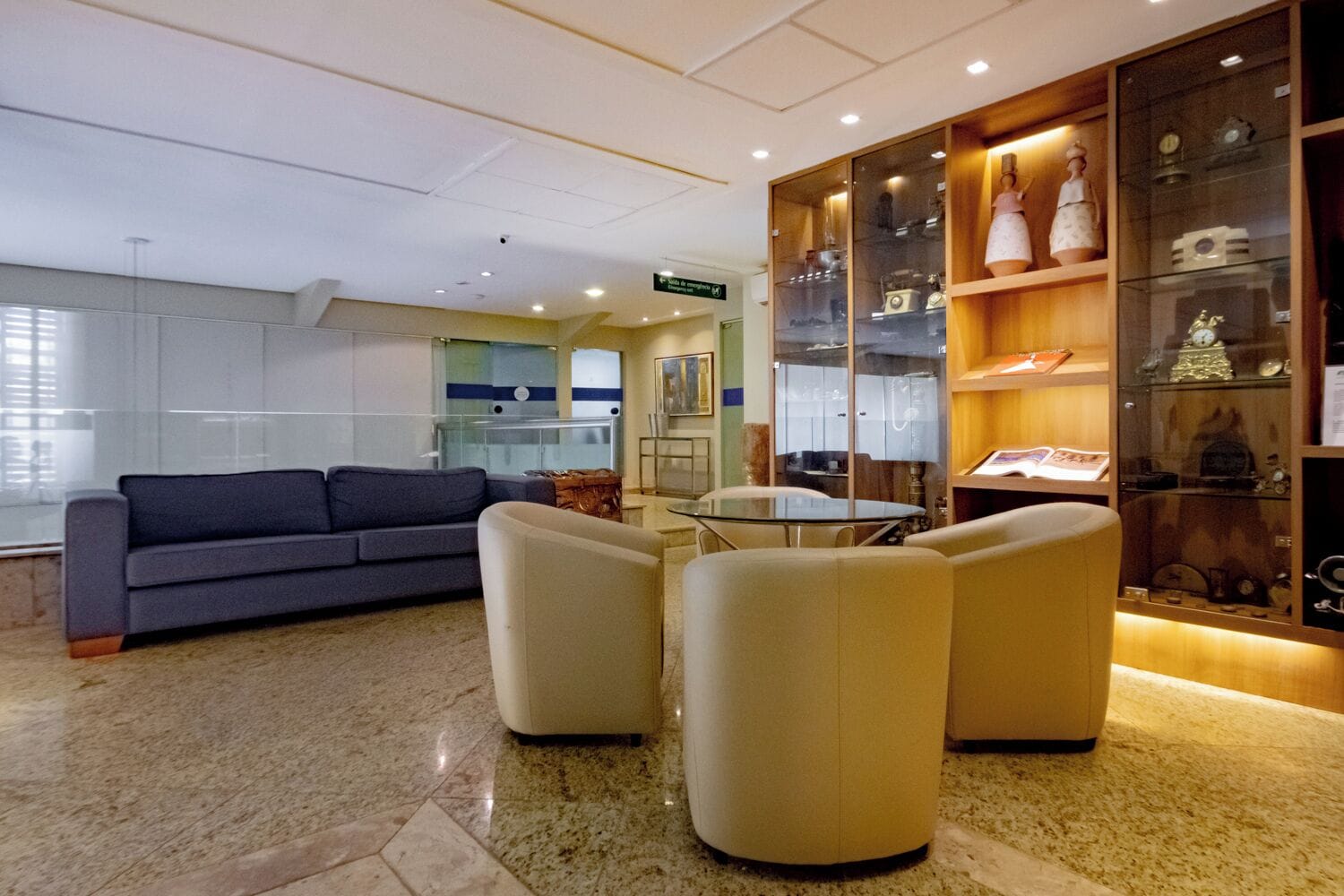 Un vestíbulo moderno con un sofá, dos sillones, una mesa de vidrio y una vitrina con elementos decorativos.