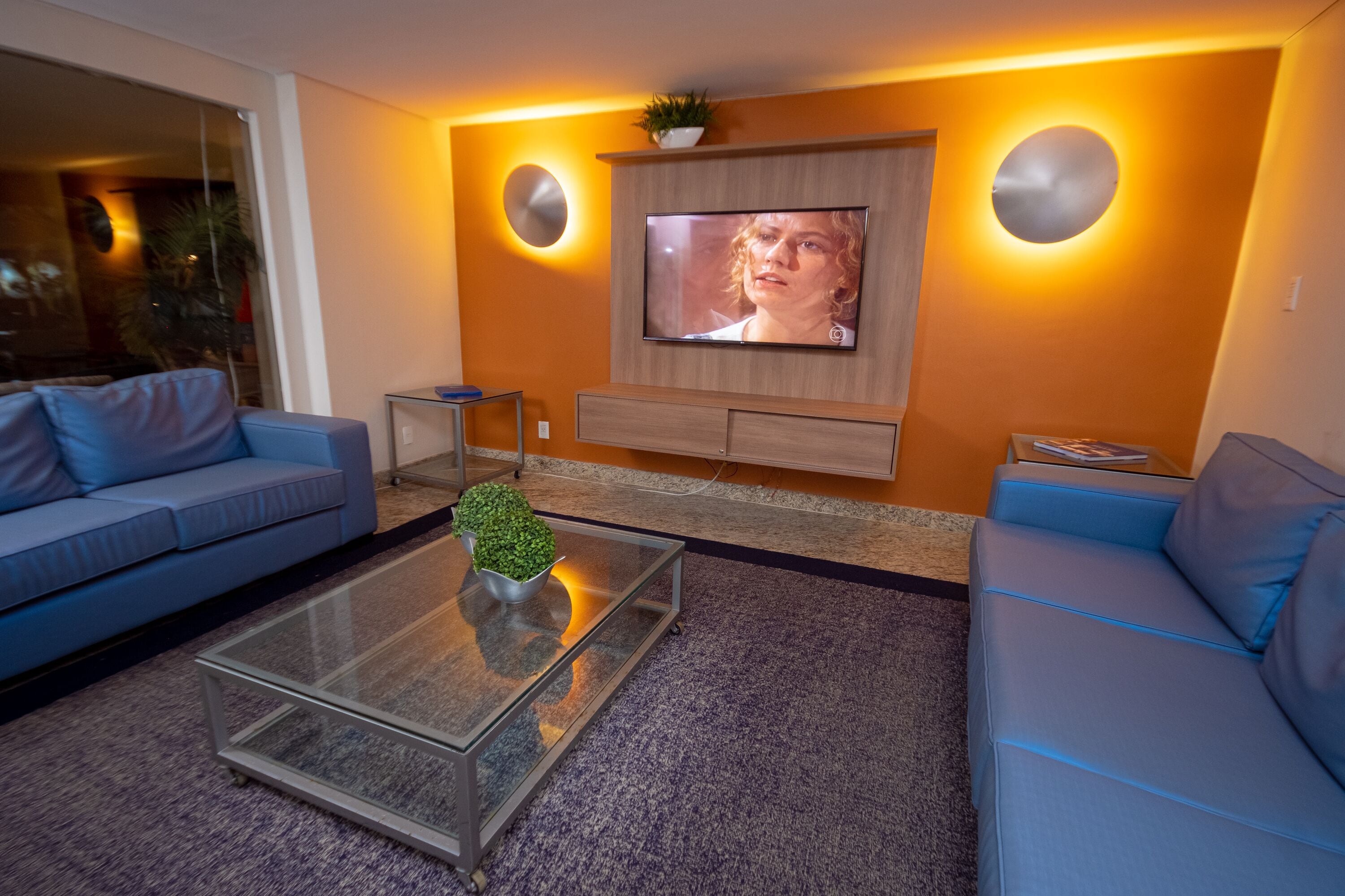 A modern living room with a flat-screen TV mounted on the wall, a glass coffee table with a plant, and two blue sofas.