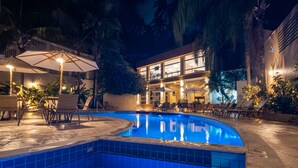 Una piscina al aire libre con sillones de descanso y sombrillas, rodeada de palmeras y un edificio con ventanas grandes.