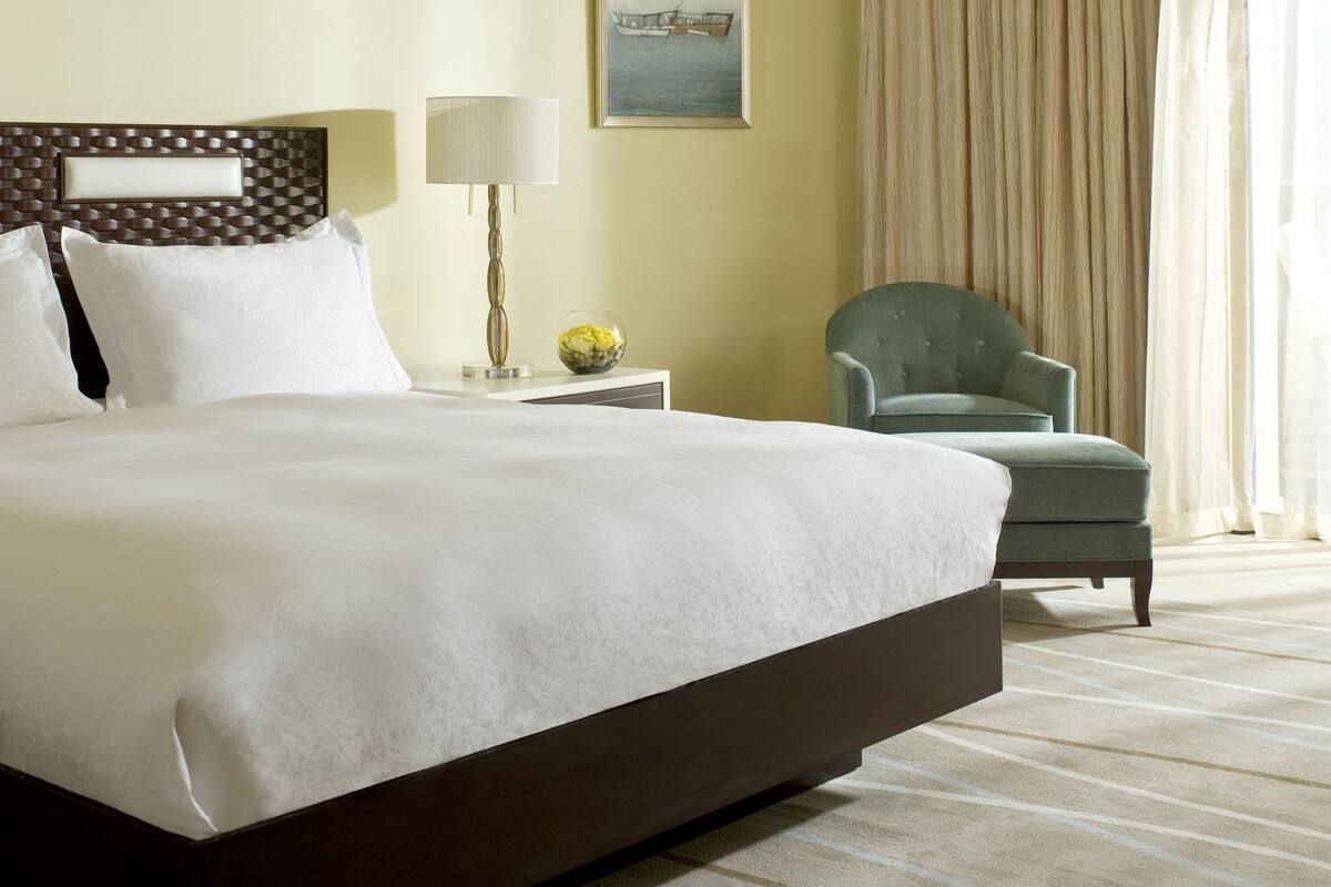 Quarto de hotel com uma cama grande, uma mesinha de cabeceira com abajur, uma cadeira e um quadro na parede.