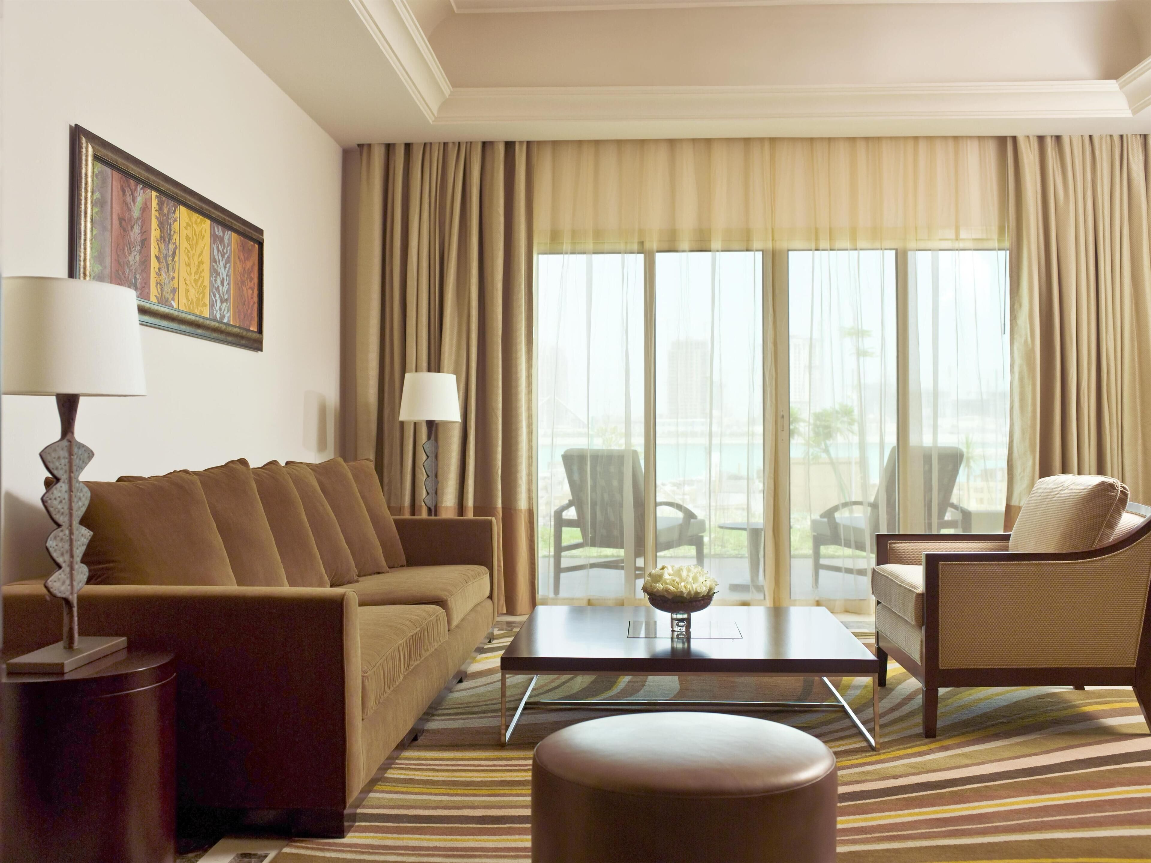 A hotel room with a sofa, armchair, coffee table, and a view of the outdoors.