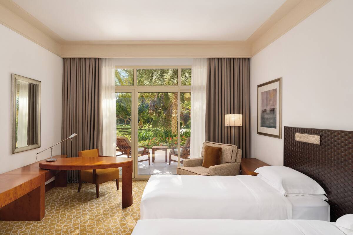 Quarto de hotel com cama, escrivaninha, cadeira, mesa e vista para um jardim através de grandes janelas.