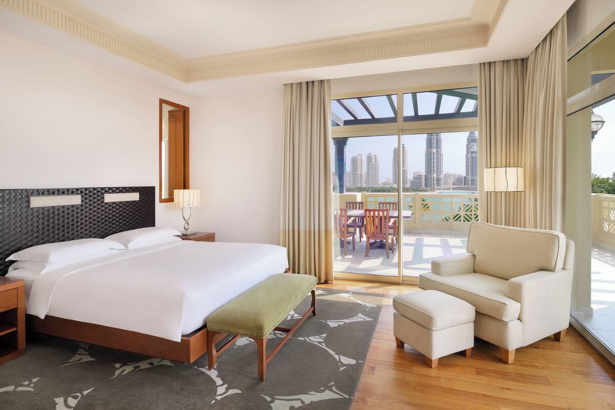 Quarto de hotel com cama grande, sofá, banqueta e varanda com vista para a cidade.