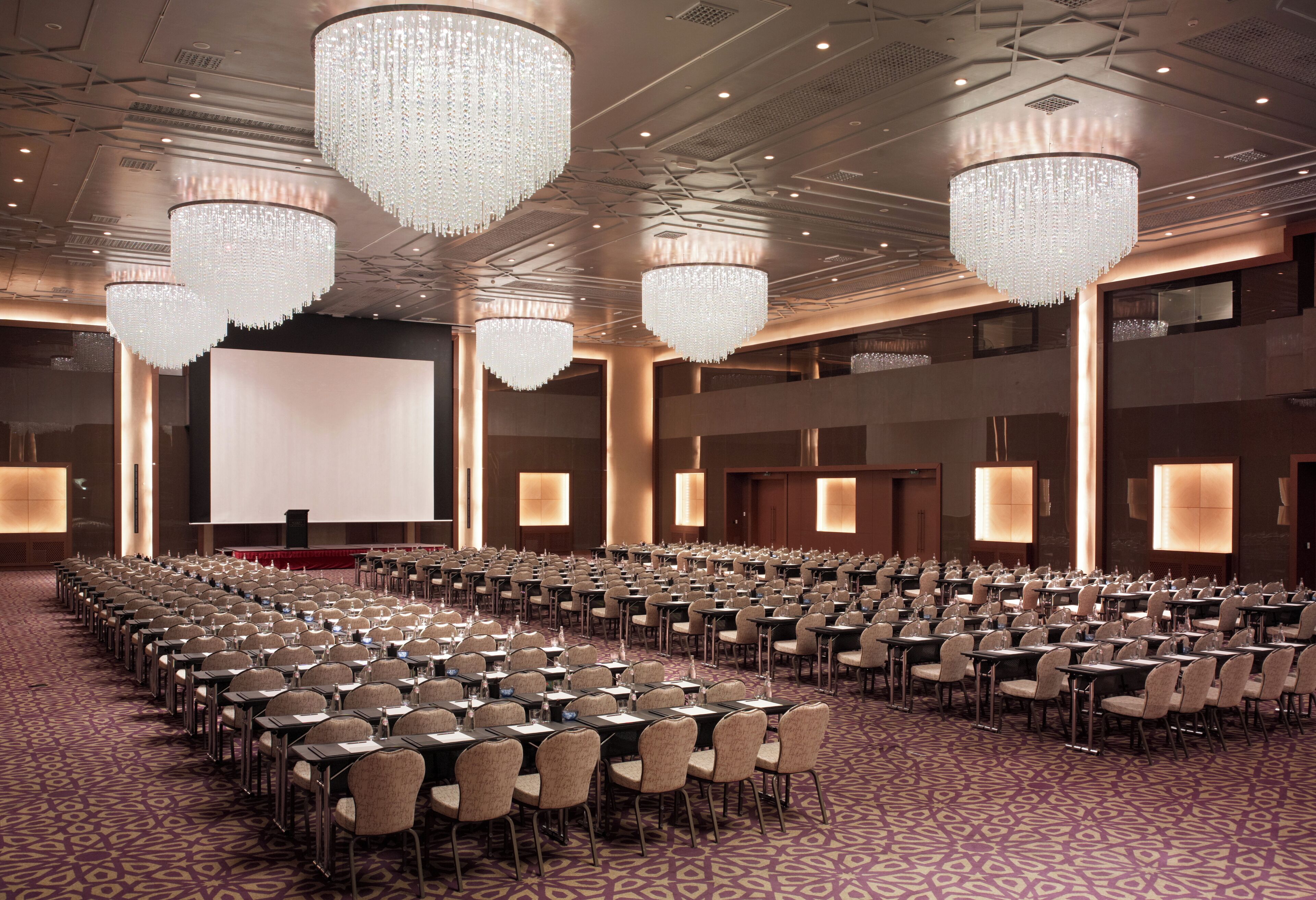 A conference room with rows of tables and chairs, large chandeliers, and a projection screen.