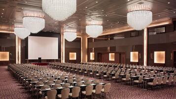A conference room with rows of tables and chairs, large chandeliers, and a projection screen.