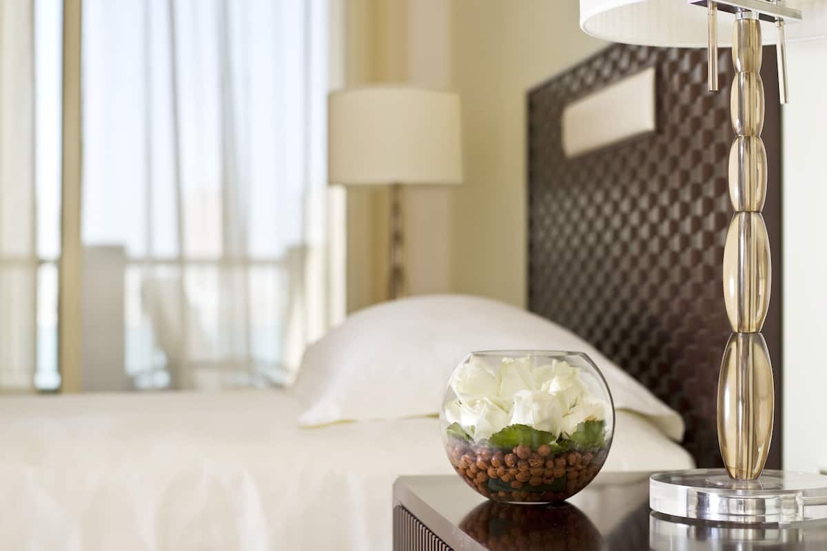 Quarto de hotel com cama, abajur e uma tigela decorativa com flores e frutos secos na mesinha de cabeceira.