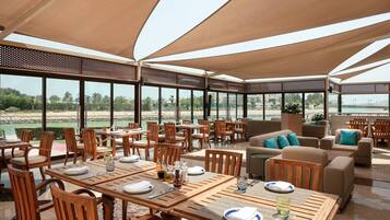 A spacious outdoor dining area with wooden tables and chairs, large windows offering a view of the landscape, and a covered seating area with sofas and cushions.