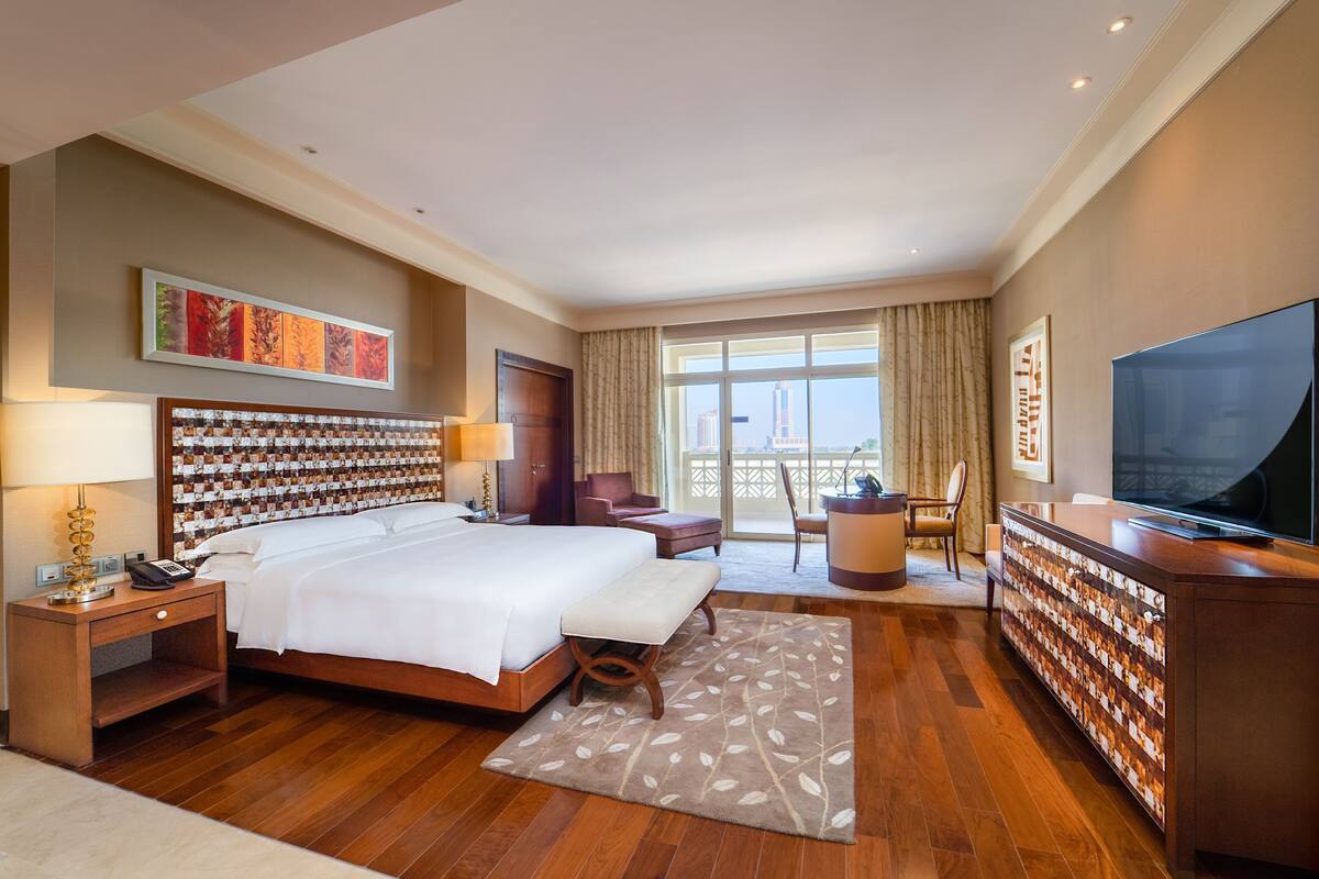 Quarto de hotel espaçoso com uma cama grande, uma mesa, televisão e varanda com vista para a cidade.