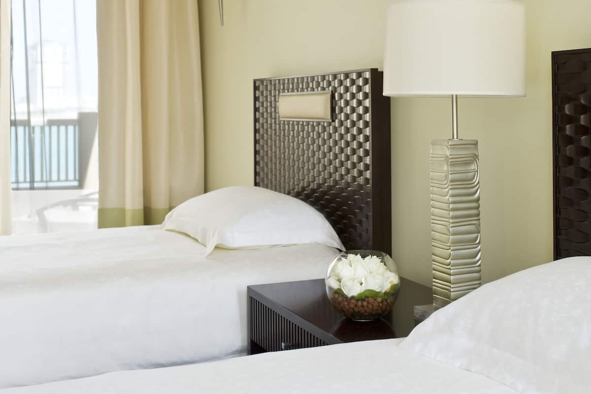 Quarto de hotel com duas camas, uma mesa de cabeceira com abajur, um vaso decorativo e uma janela com cortinas.