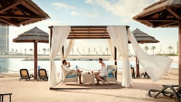 A beach cabana with a thatched roof, a covered seating area, and a view of the sea and palm trees in the background.