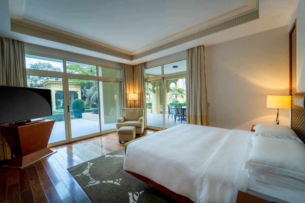 Quarto de hotel espaçoso com uma cama grande, televisão de tela plana, área de estar com uma cadeira e mesa, e uma varanda com vista para o jardim.
