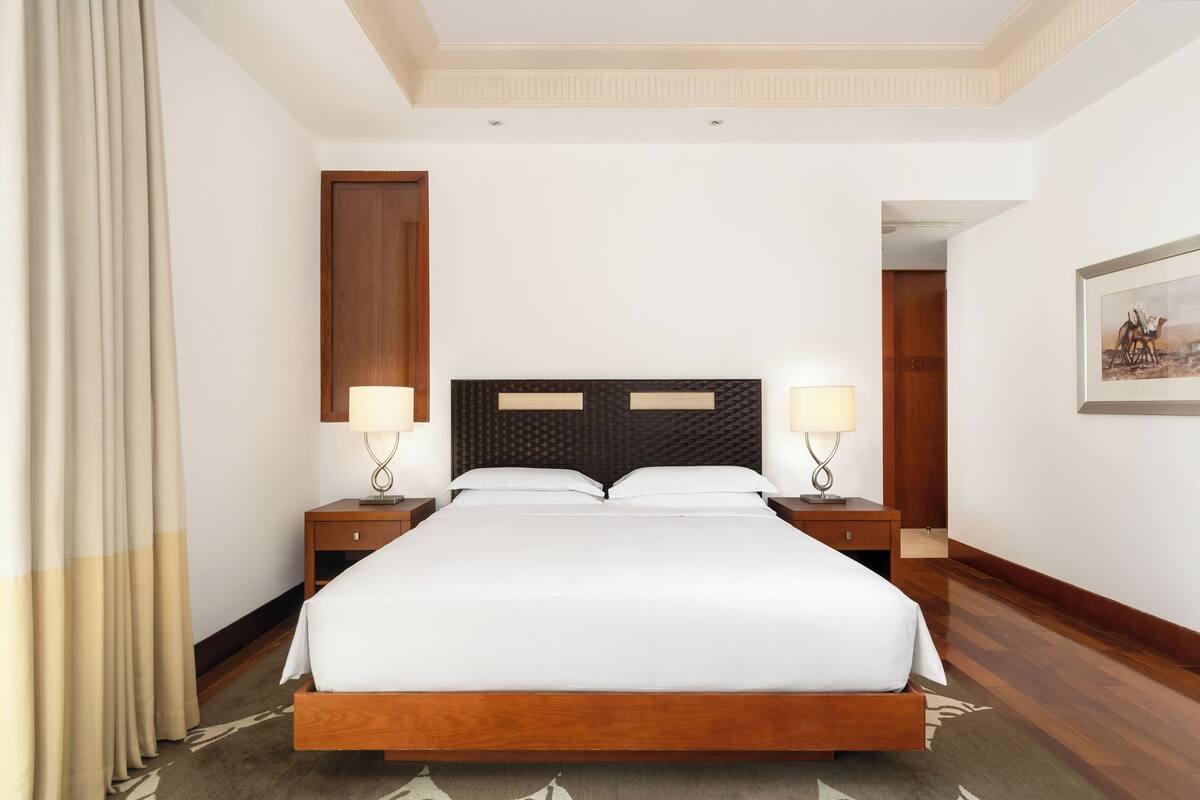 Quarto de hotel com uma cama grande, cabeceira de madeira, duas mesas de cabeceira com abajures, uma porta e um quadro na parede.