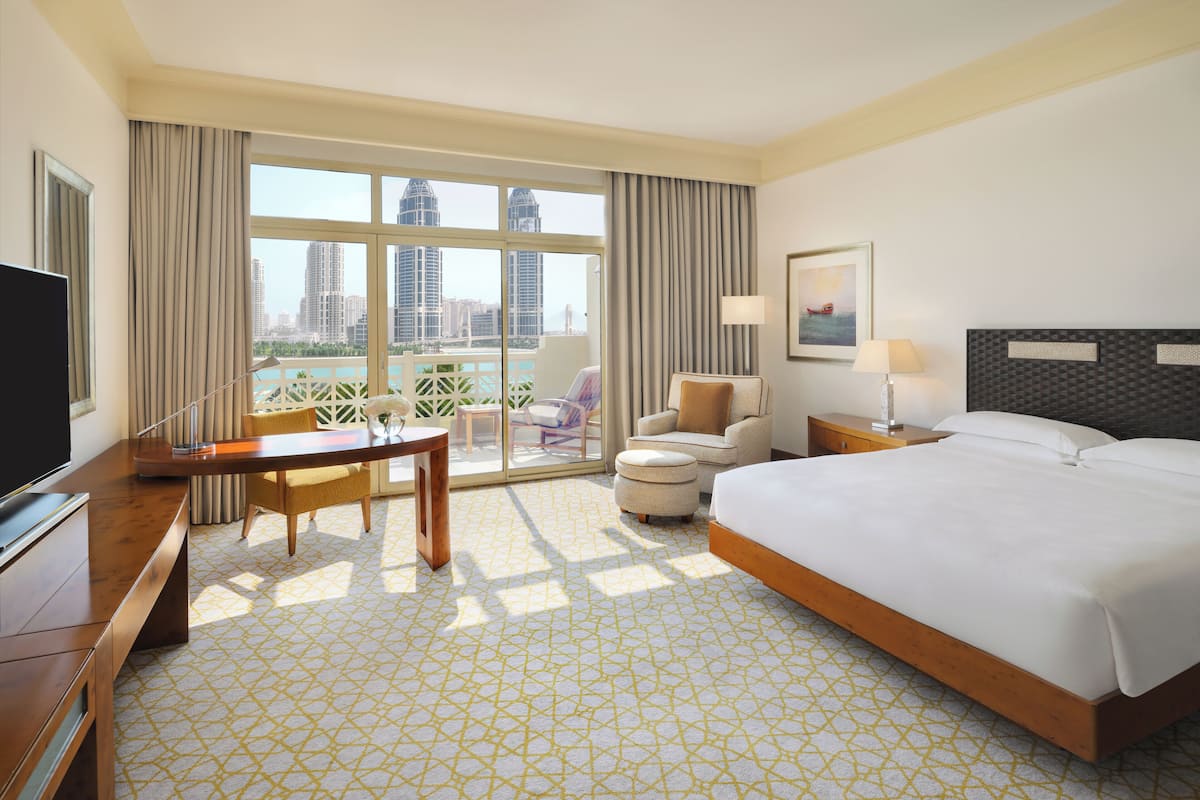 Quarto de hotel com uma cama grande, uma mesa, uma cadeira, uma televisão e uma varanda com vista para edifícios altos.