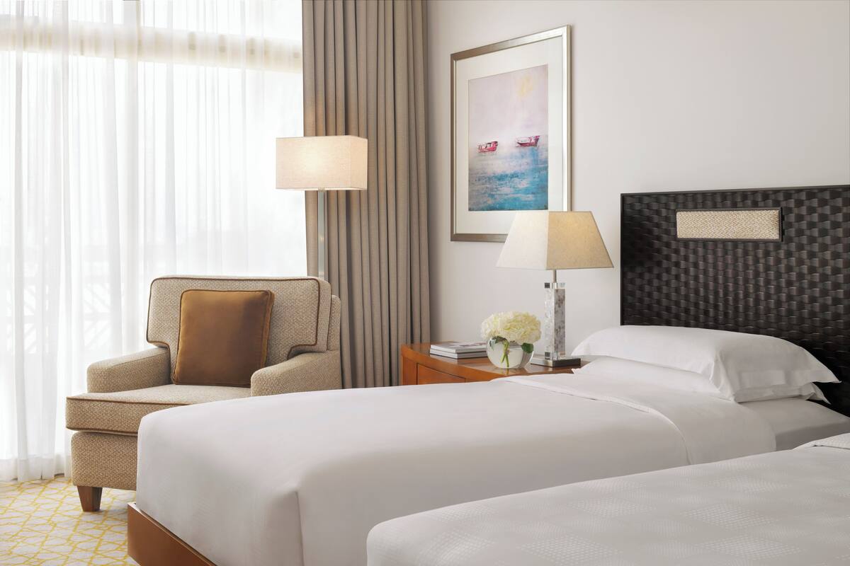 Quarto de hotel com duas camas, uma cadeira, uma mesa de cabeceira com abajur e um quadro na parede.