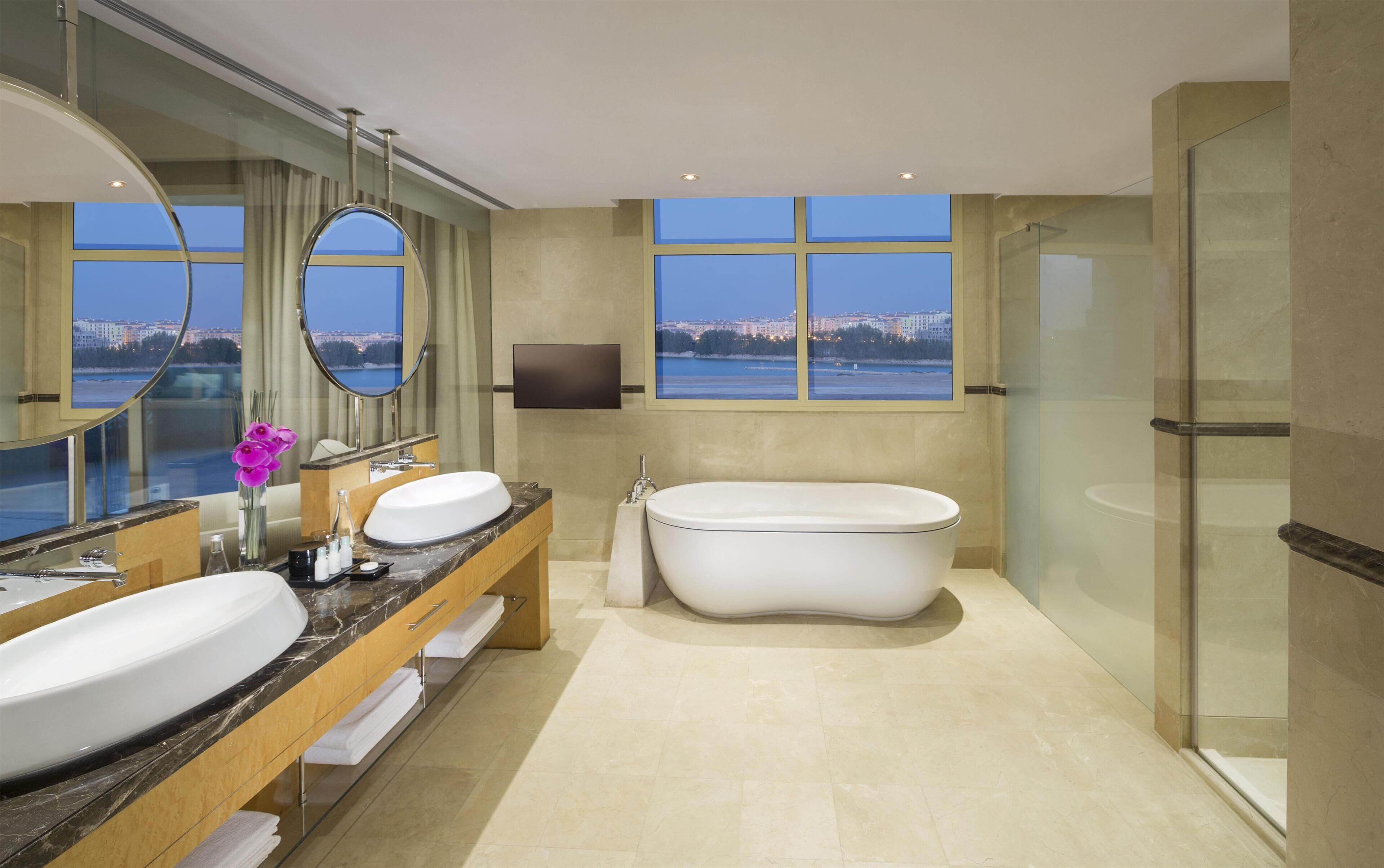 A modern bathroom with a large bathtub, two sinks, a large mirror, and a view of a cityscape through the windows.