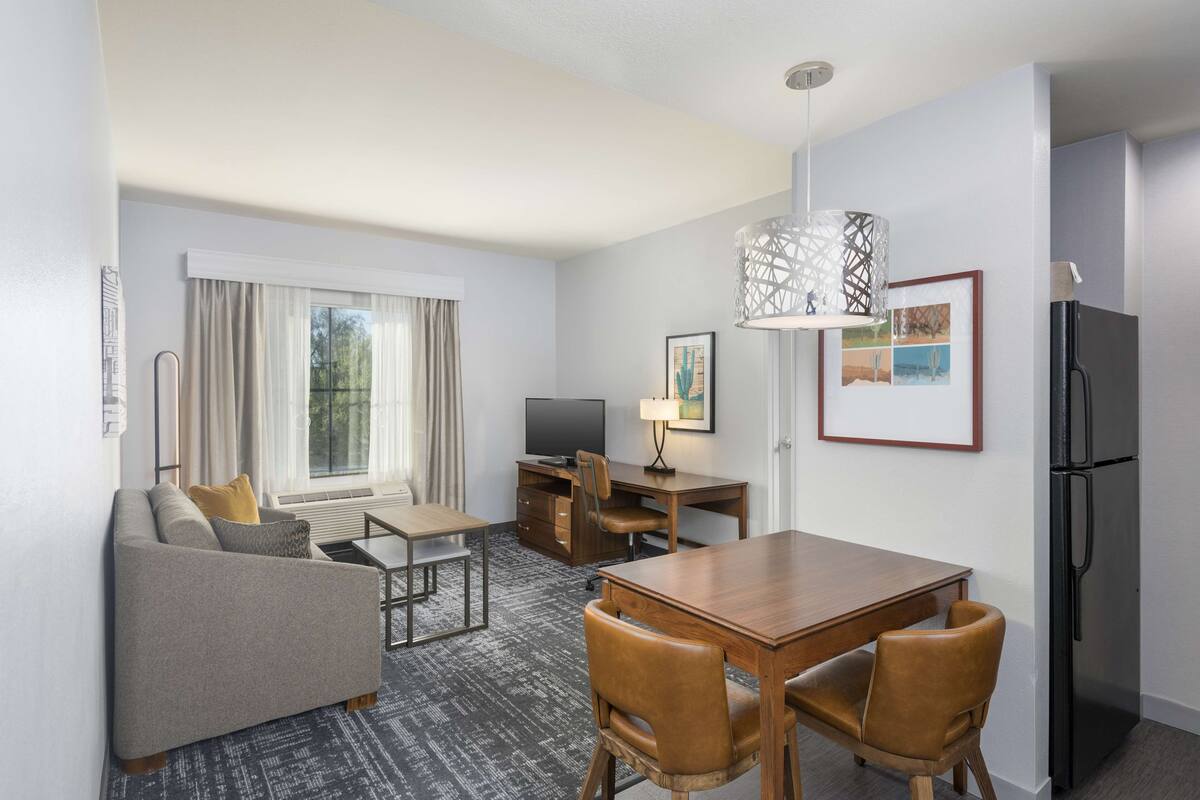 A compact hotel room with a sofa, a dining table with chairs, a TV, a refrigerator, and a window with curtains.