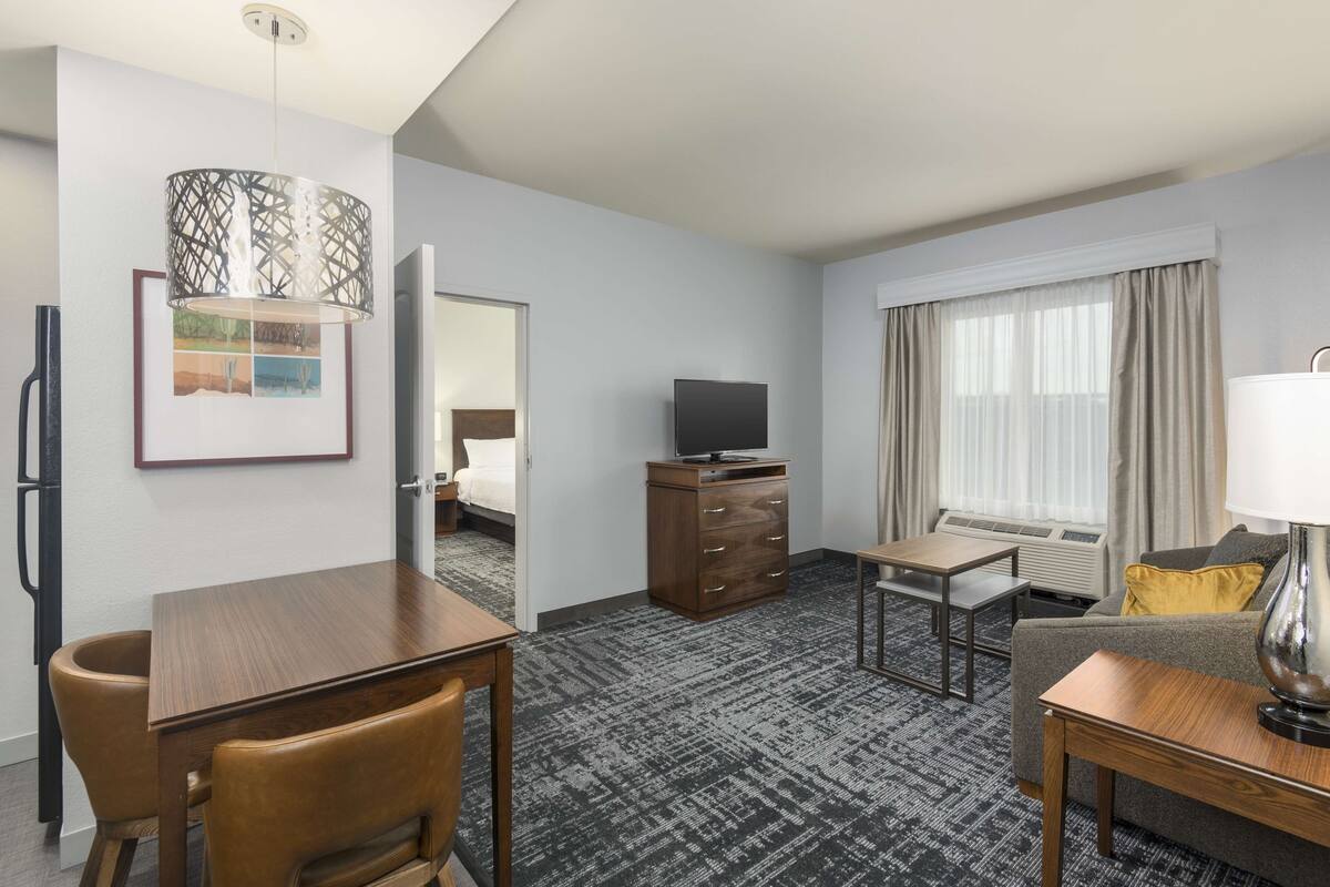 A hotel room with a wooden desk, a television, a sofa, a lamp, and a large window with curtains.