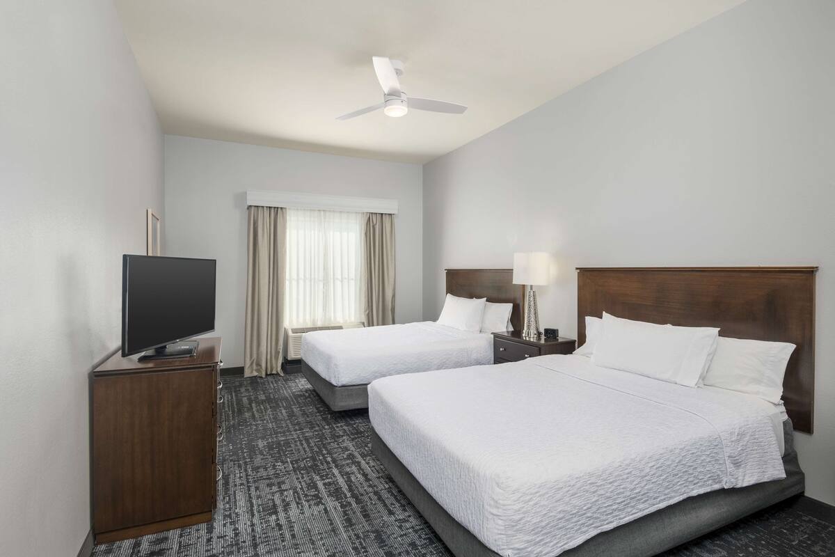 A hotel room with two beds, a TV on a cabinet, a ceiling fan, and a window with curtains.