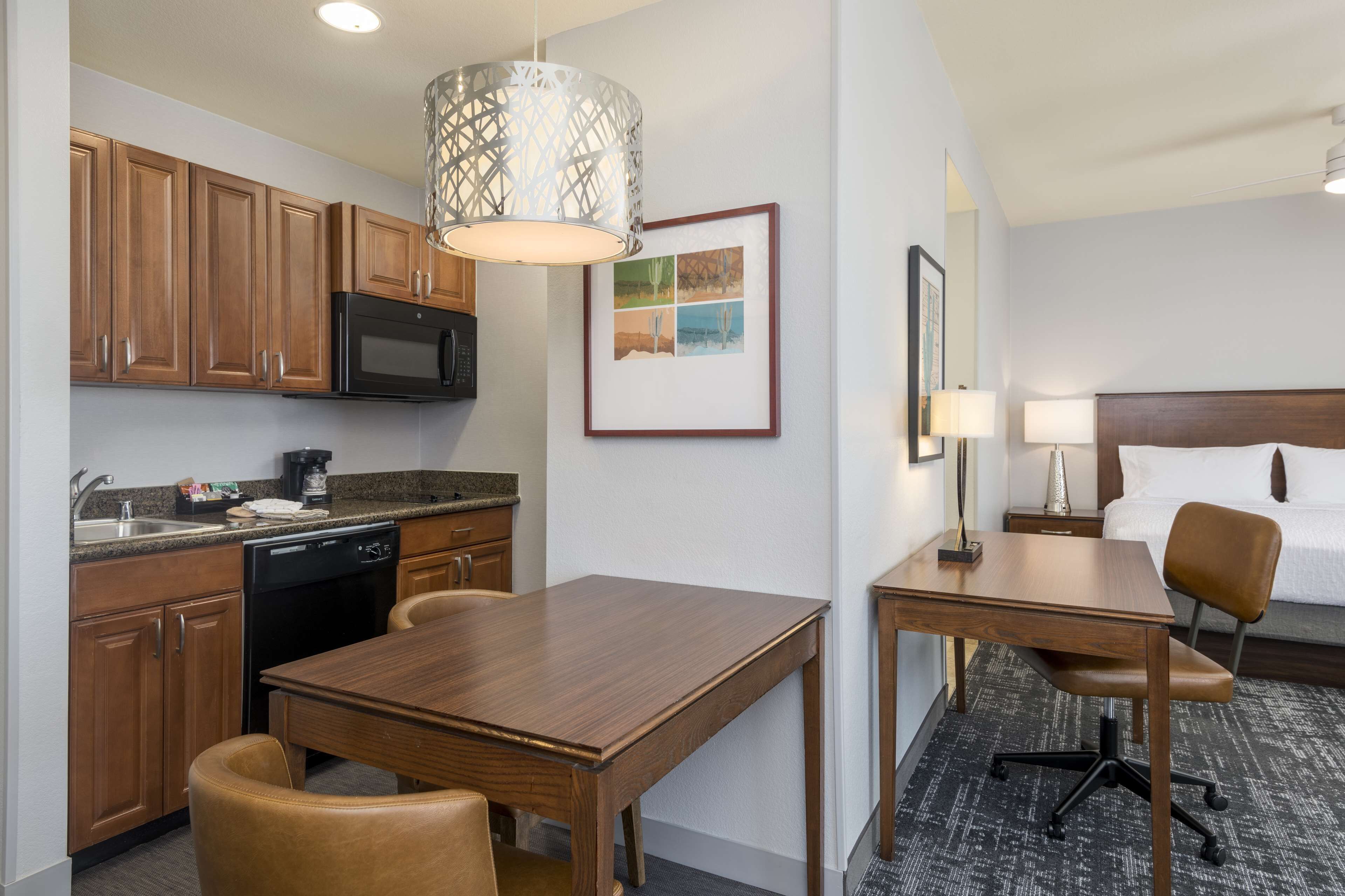A hotel room with a wooden dining table, a kitchen area with a microwave, and a bed with a headboard.