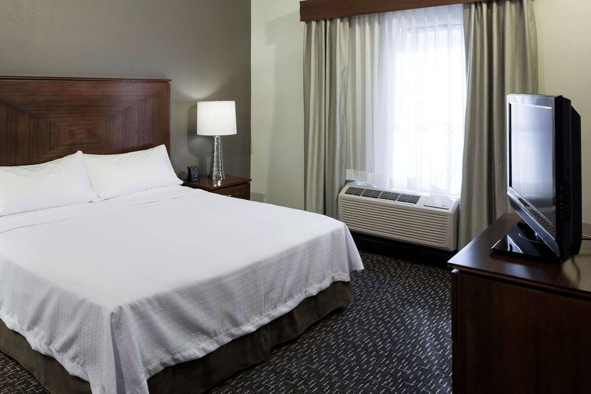 A hotel room with a large bed, a television, an air conditioning unit, a nightstand with a lamp, and a window with curtains.