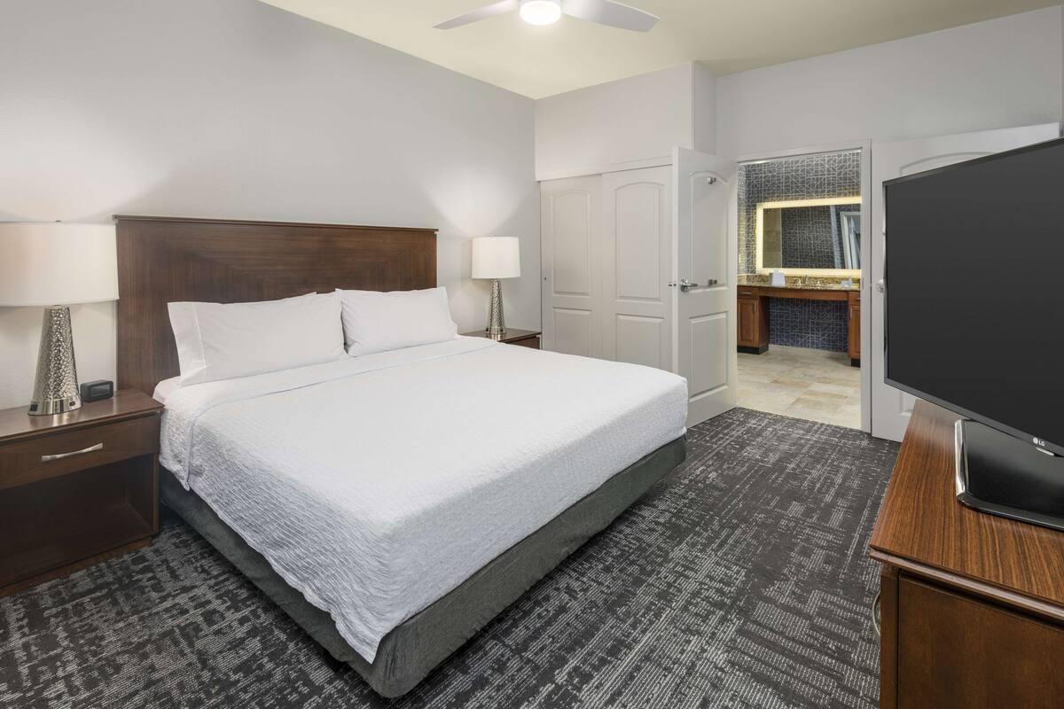 A hotel room with a large bed, a TV, a desk with a lamp, and a bathroom visible through an open door.