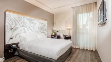 Ein Hotelzimmer mit einem großen Bett, einem Schreibtisch mit Lampe, einem Telefon und einem an der Wand befestigten Fernseher.