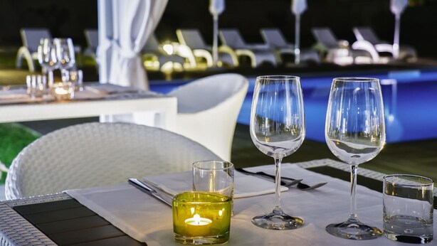 Una mesa de comedor dispuesta con copas de vino, una vela y cubiertos, con vistas a una zona de piscina con sillones de descanso.