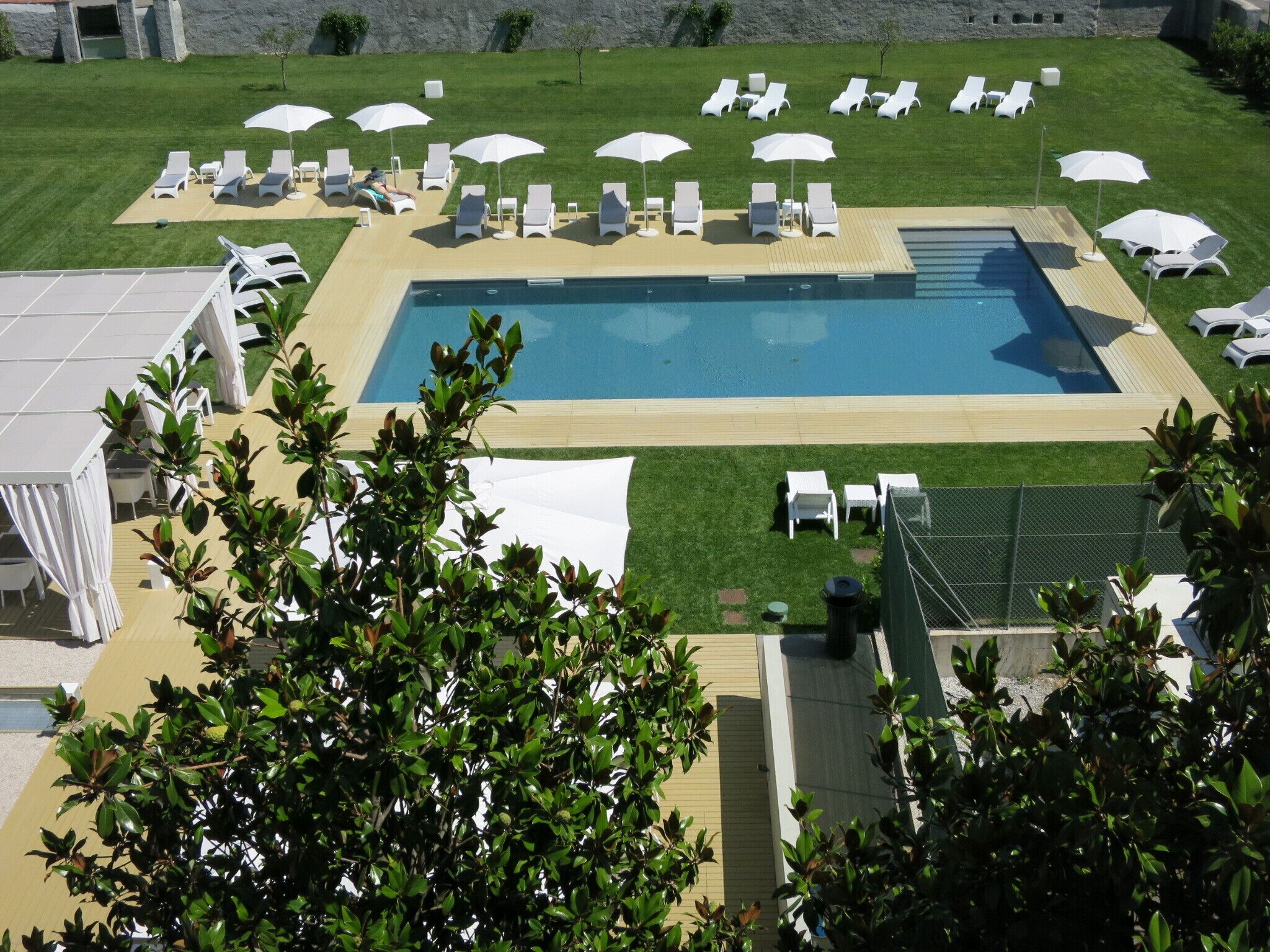Un área de piscina con sillas de descanso y sombrillas, rodeada de césped cuidado y un muro de piedra al fondo.