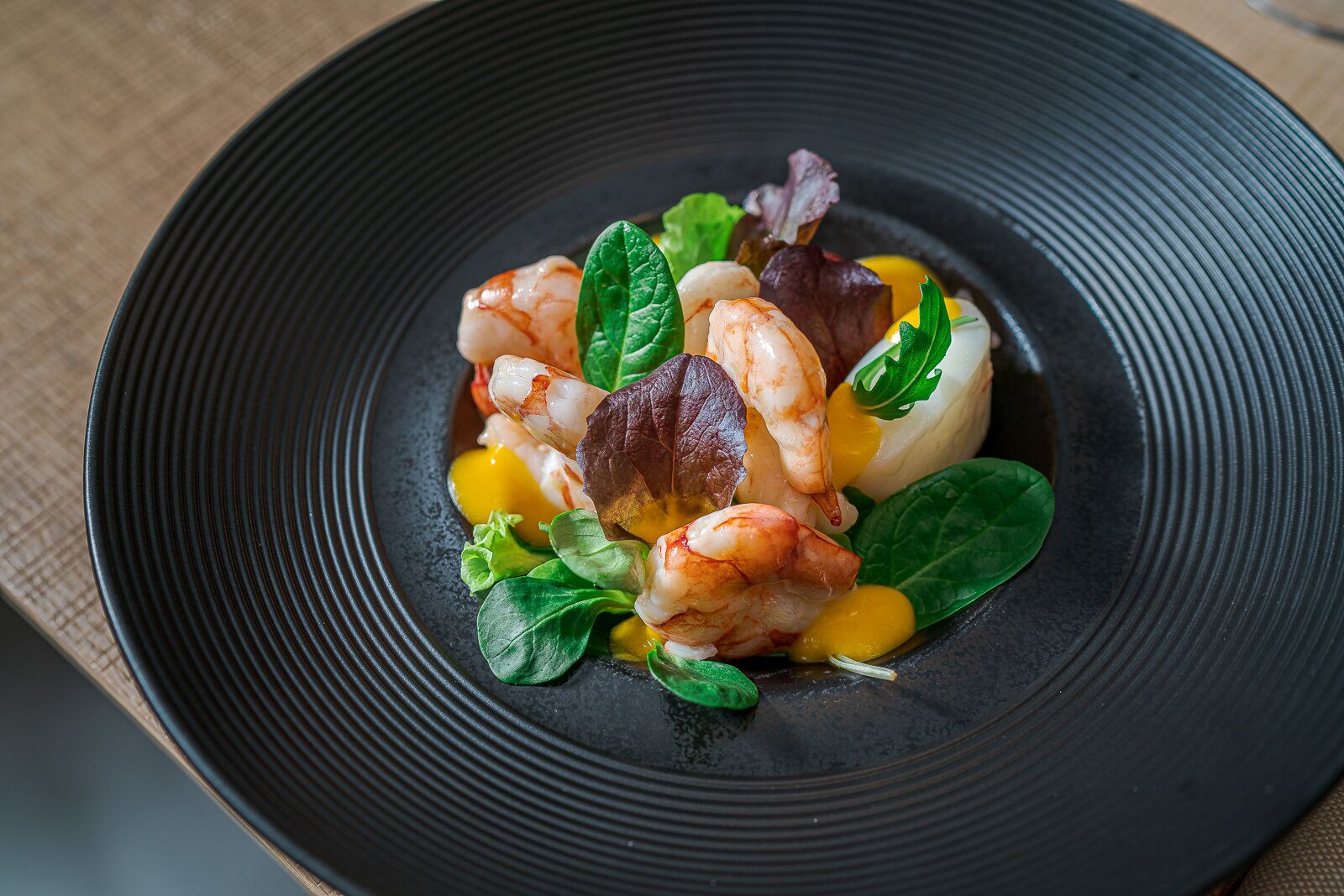 Une assiette noire contenant un plat de fruits de mer, notamment des crevettes et des légumes verts, posée sur une surface texturée.