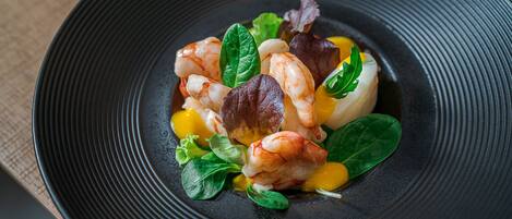 A black plate with a seafood dish, including shrimp and greens, on a textured surface.